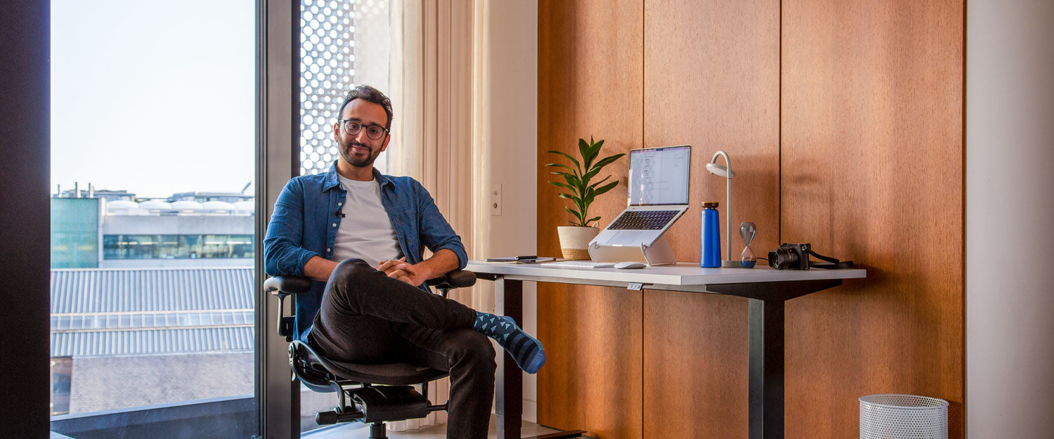 Kuva podcastaaja Ali Abdaalista istumassa grafiitinvärisessä Herman Miller Aeron -työtuolissa Nevi-seisomapöydän edessä, jossa on mustat jalat ja valkoinen pöytälevy, sekä kannettava tietokone Oripura-kannettavan telineessä
