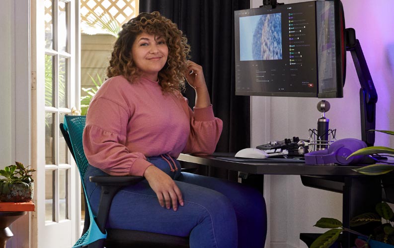 Ammattilaisstriimaaja ja ääninäyttelijä Friskk istuu Herman Miller Sayl -pelituolilla, joka on syvän sininen ja musta, mustalla Nevi-istuma-seisomapöydällä ja Ollin-näytönjalustalla.