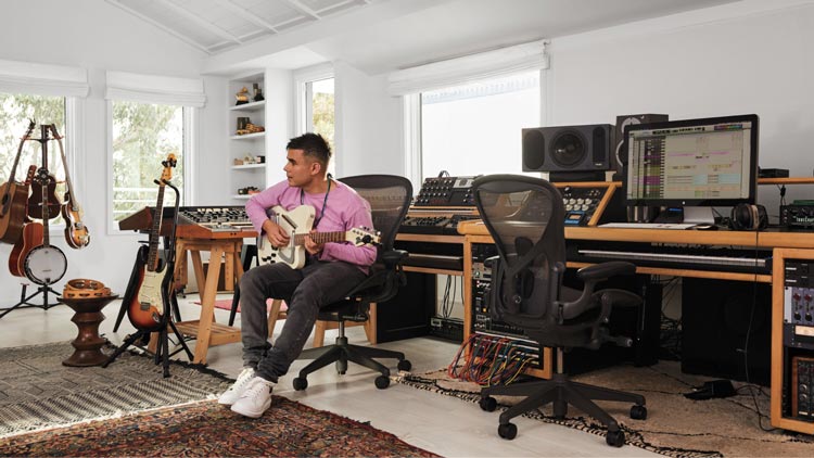 Valokuva muusikosta, tuottajasta ja säveltäjästä Rostamista, joka soittaa kitaraa musiikkistudiossaan istuen grafiitinvärisessä Aeron-työtuolissa