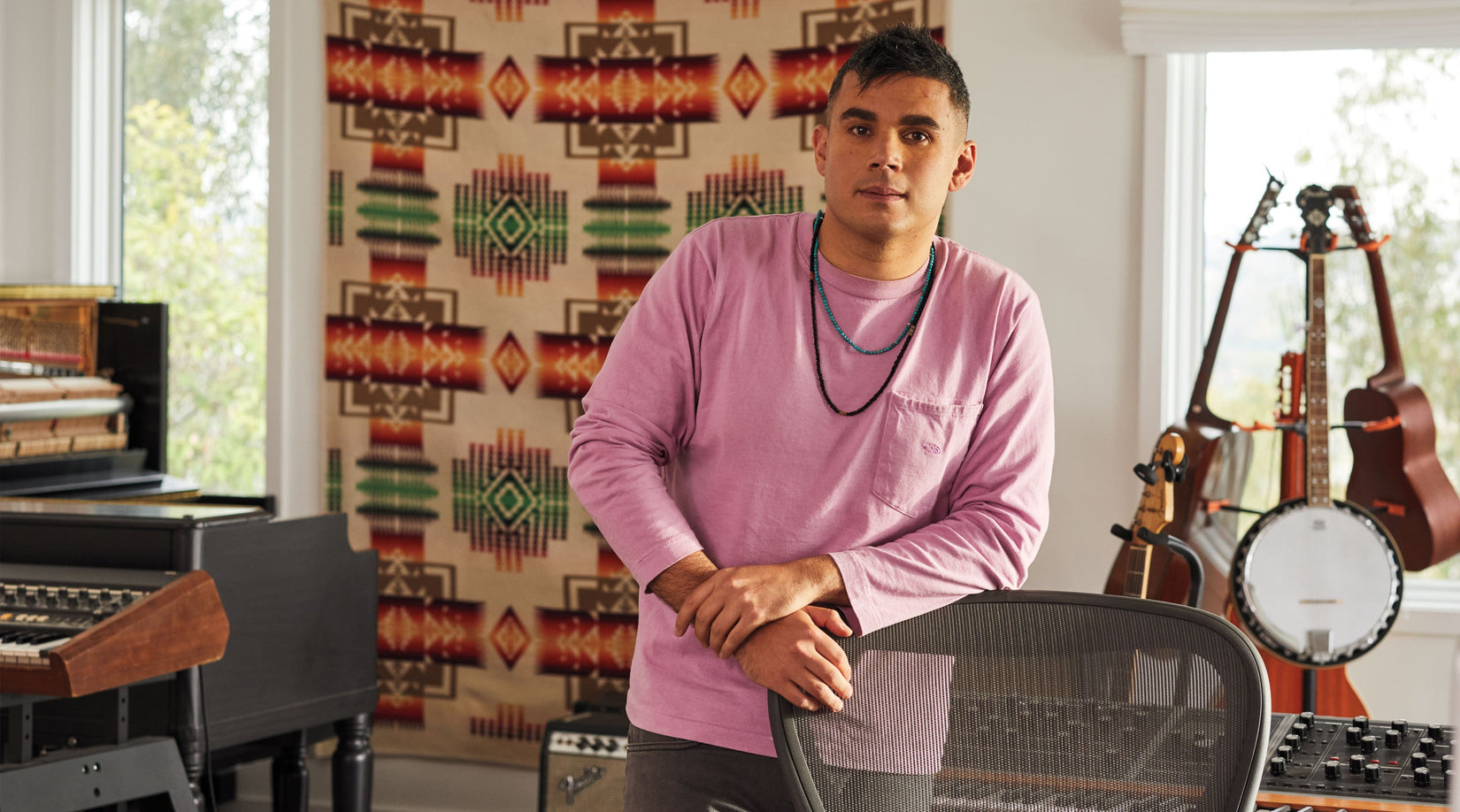 Rostam, muusikko, tuottaja ja säveltäjä, seisoo nojautuneena grafiittiväriseen Aeron-työtuoliin, taustalla musiikki-instrumentteja.