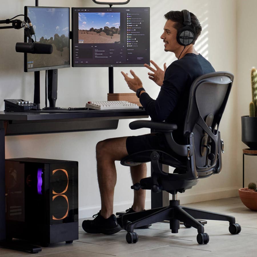 Pelaaja ja striimaaja Xaryu pelikokoonpanossa istumassa Herman Miller Aeron -pelituolilla, mustalla Nevi-seisomapöydällä ja kahdella monitorilla Ollin-monitorivarsilla