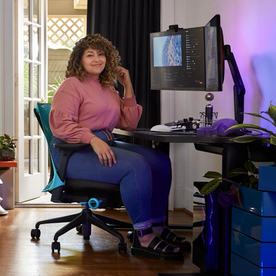 Ammattilaisstriimaaja ja ääninäyttelijä Friskk istuu Herman Miller Sayl -pelituolissa, väri Ocean deep blue ja musta, mustalla Nevi-seisomapöydällä, jossa on näyttö Oliin-näytönjalustalla.